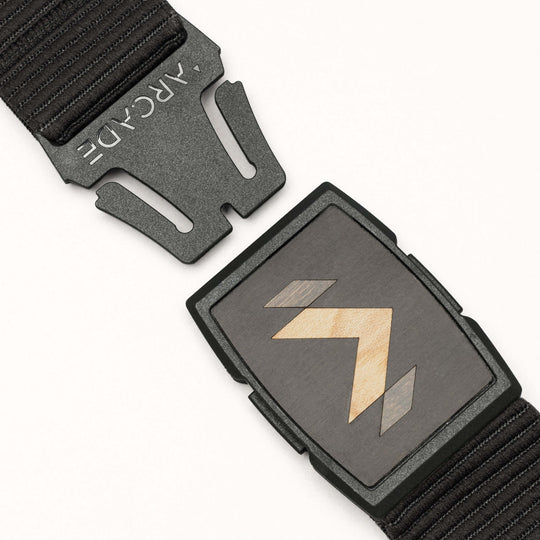 A close-up of the Carver stretch belt by Arcade Belts, featuring a rectangular buckle with a geometric arrow design. The belt is partially unclasped, displaying the ARCADE logo on the clasp.