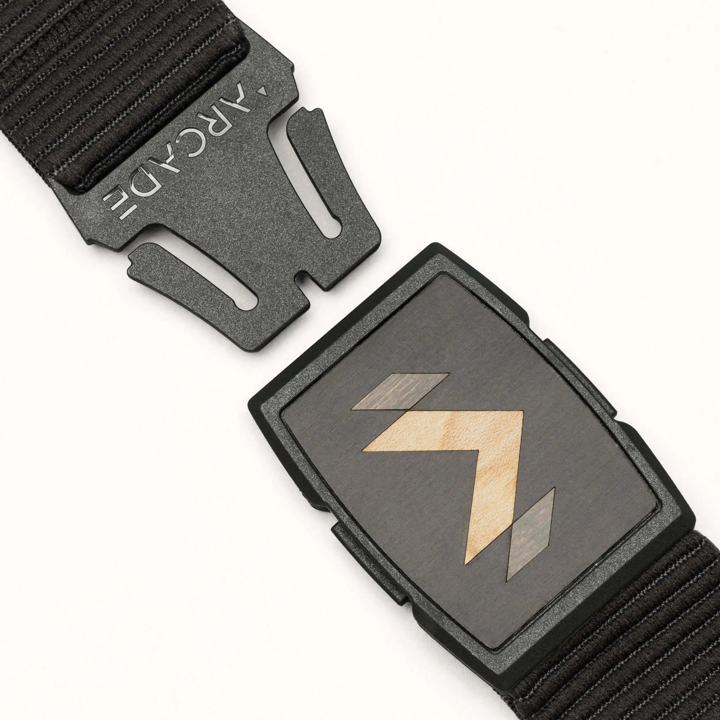 A close-up of the Carver stretch belt by Arcade Belts, featuring a rectangular buckle with a geometric arrow design. The belt is partially unclasped, displaying the ARCADE logo on the clasp.