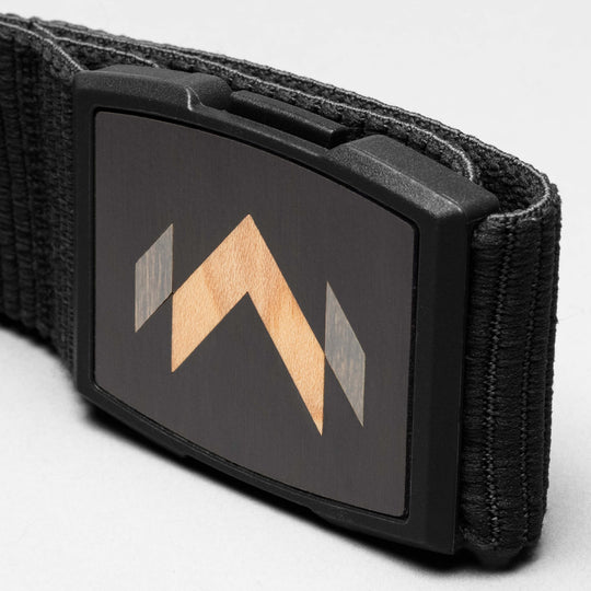 A close-up of the Arcade Belts Carver shows its rectangular buckle with a wooden inlay arrow design on a dark background. The textured fabric showcases the Carver stretch belt’s unique style.