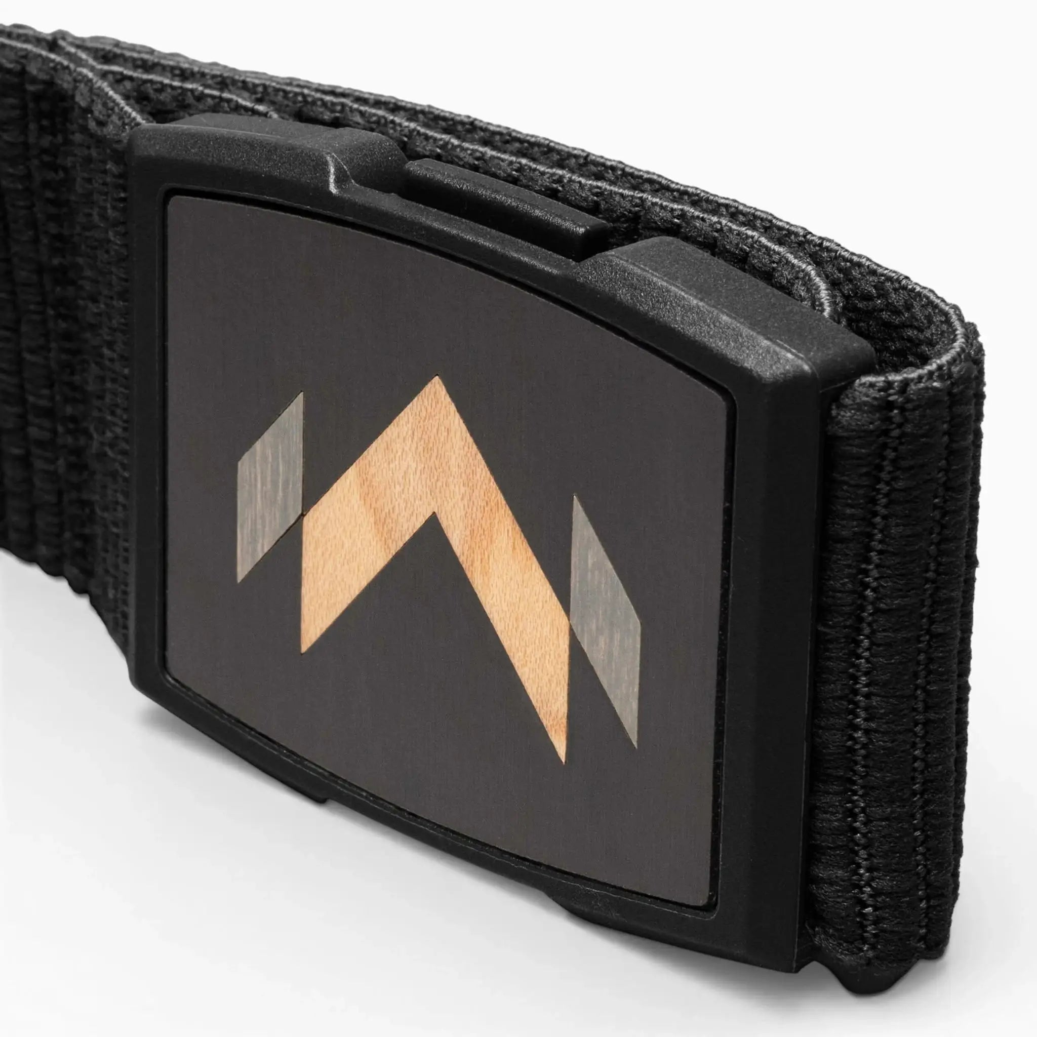 A close-up of the Arcade Belts Carver shows its rectangular buckle with a wooden inlay arrow design on a dark background. The textured fabric showcases the Carver stretch belt’s unique style.