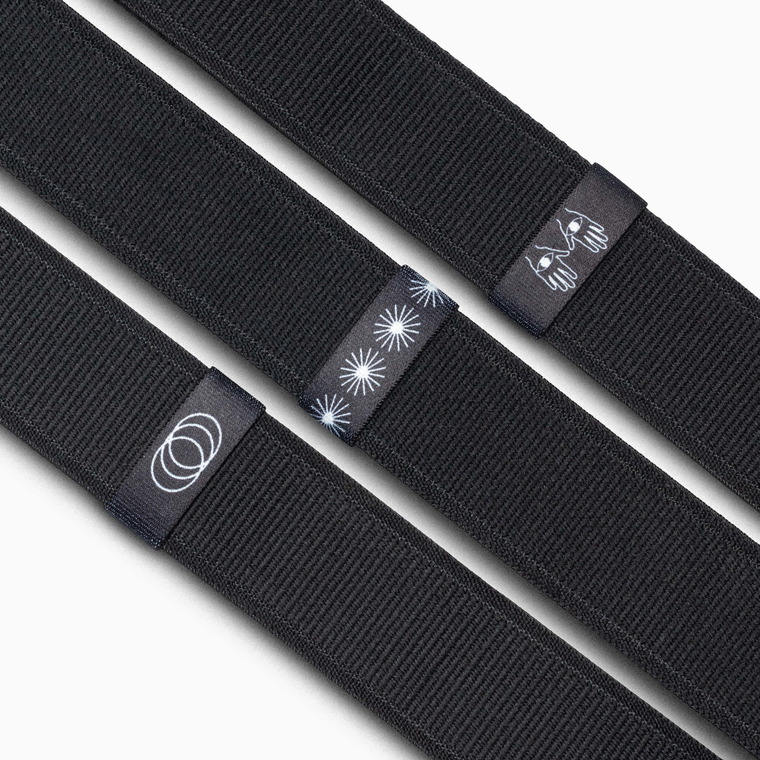 Three black fabric Arcade Belts Belt Loopkeepers are shown side by side, each with a unique black tag featuring white designs: concentric circles, starbursts, and a stylized bee. Ideal as replacement elastic loops for Standard Width Adventure Belts.