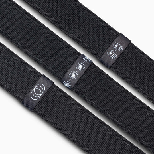 Three black fabric Arcade Belts Belt Loopkeepers are shown side by side, each with a unique black tag featuring white designs: concentric circles, starbursts, and a stylized bee. Ideal as replacement elastic loops for Standard Width Adventure Belts.