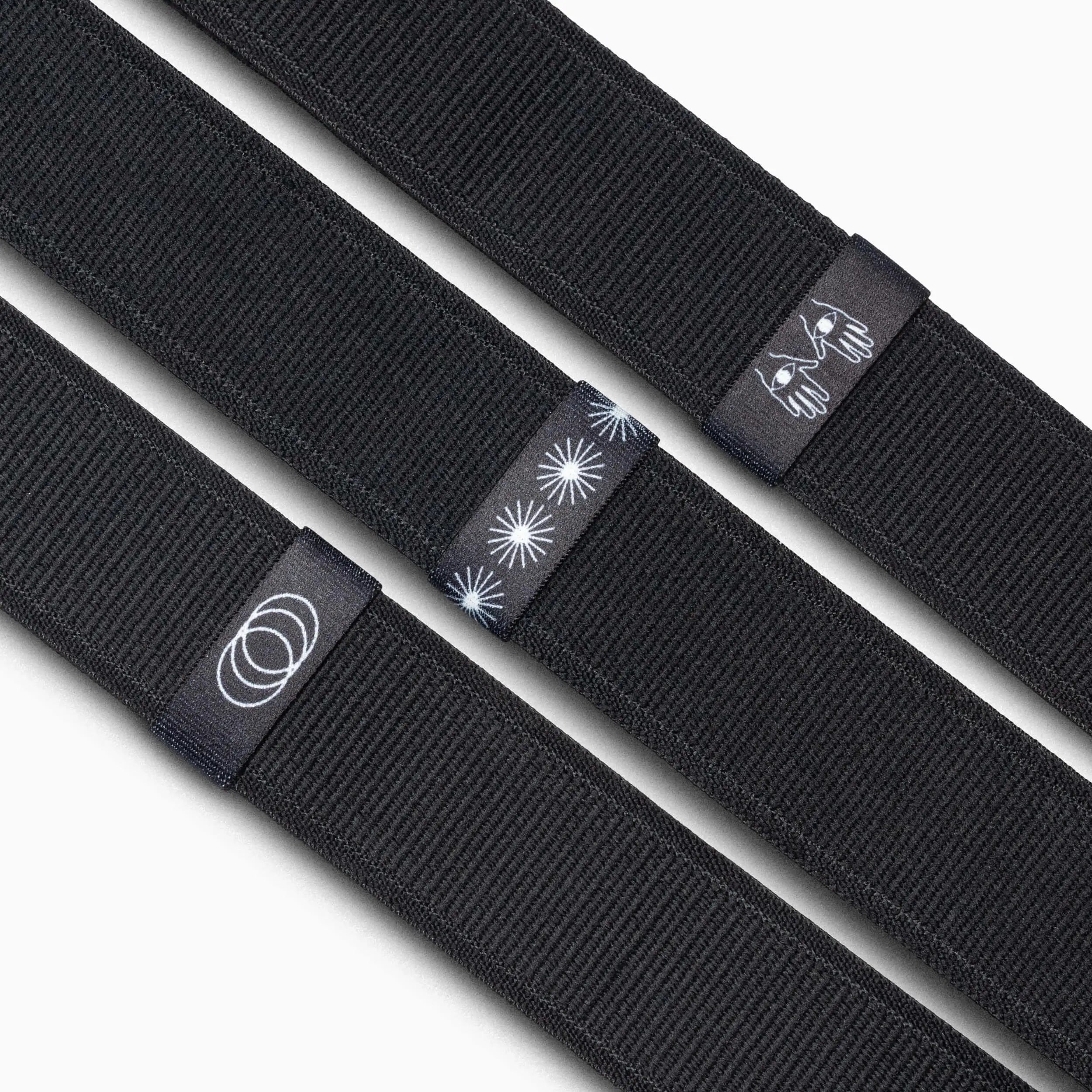 Three black fabric Arcade Belts Belt Loopkeepers are shown side by side, each with a unique black tag featuring white designs: concentric circles, starbursts, and a stylized bee. Ideal as replacement elastic loops for Standard Width Adventure Belts.