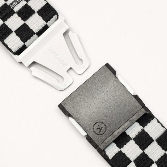 Close-up of the Arcade Belts "Check" belt, featuring black and white checkered fabric, a white plastic buckle with clasp and sad face icon. Its performance stretch design ensures comfort and security against a light background.