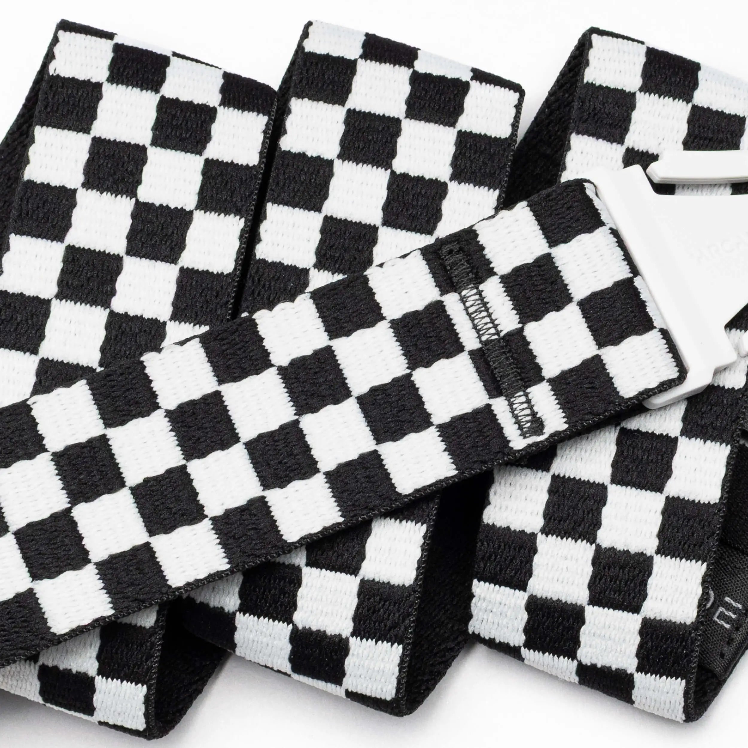 A close-up of the Arcade Belts Check strap shows its black-and-white checkered design and silver buckle. The performance stretch belt is folded and layered against a white background, emphasizing its comfort and flexibility for movement.