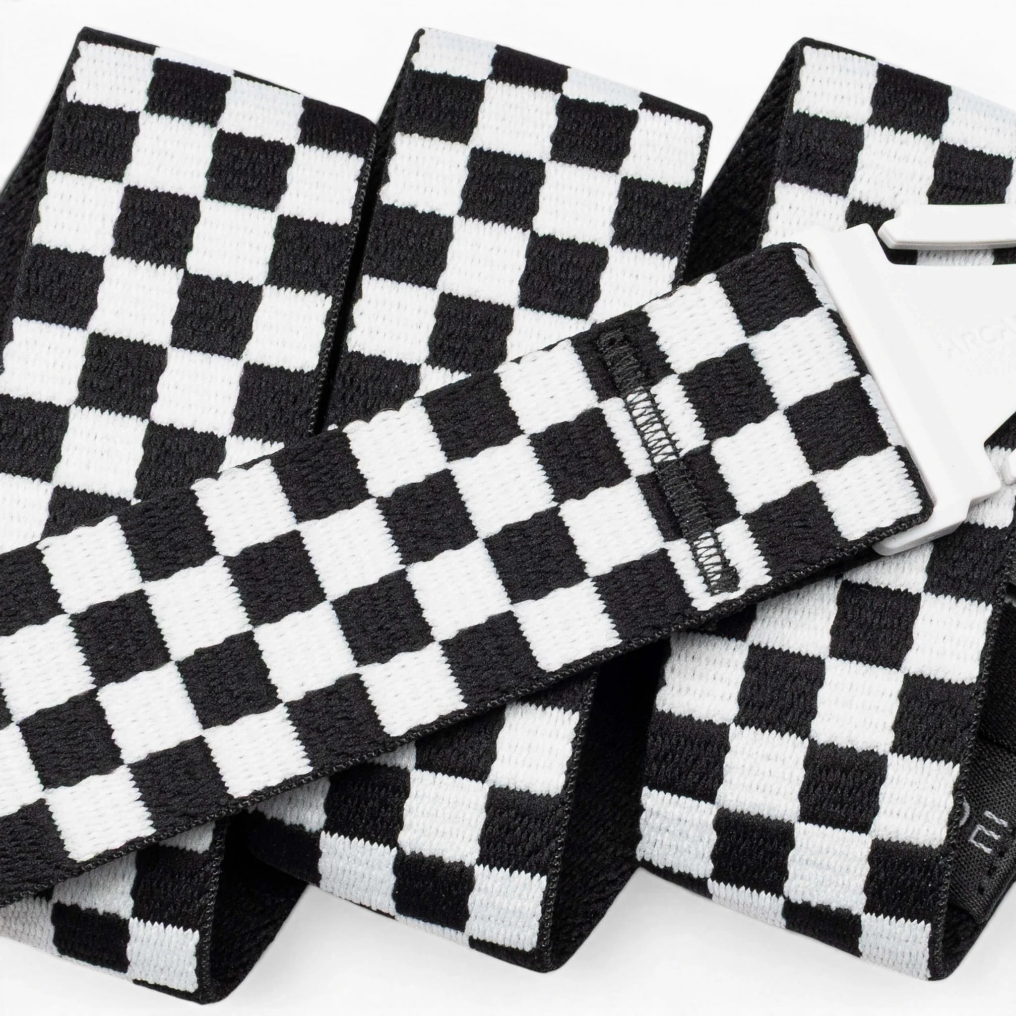 A close-up of the Arcade Belts Check strap shows its black-and-white checkered design and silver buckle. The performance stretch belt is folded and layered against a white background, emphasizing its comfort and flexibility for movement.