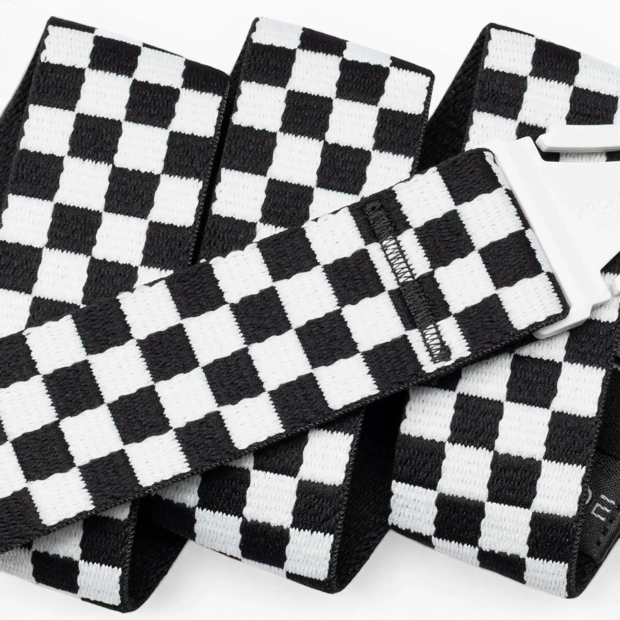 A close-up of the Arcade Belts Check strap shows its black-and-white checkered design and silver buckle. The performance stretch belt is folded and layered against a white background, emphasizing its comfort and flexibility for movement.