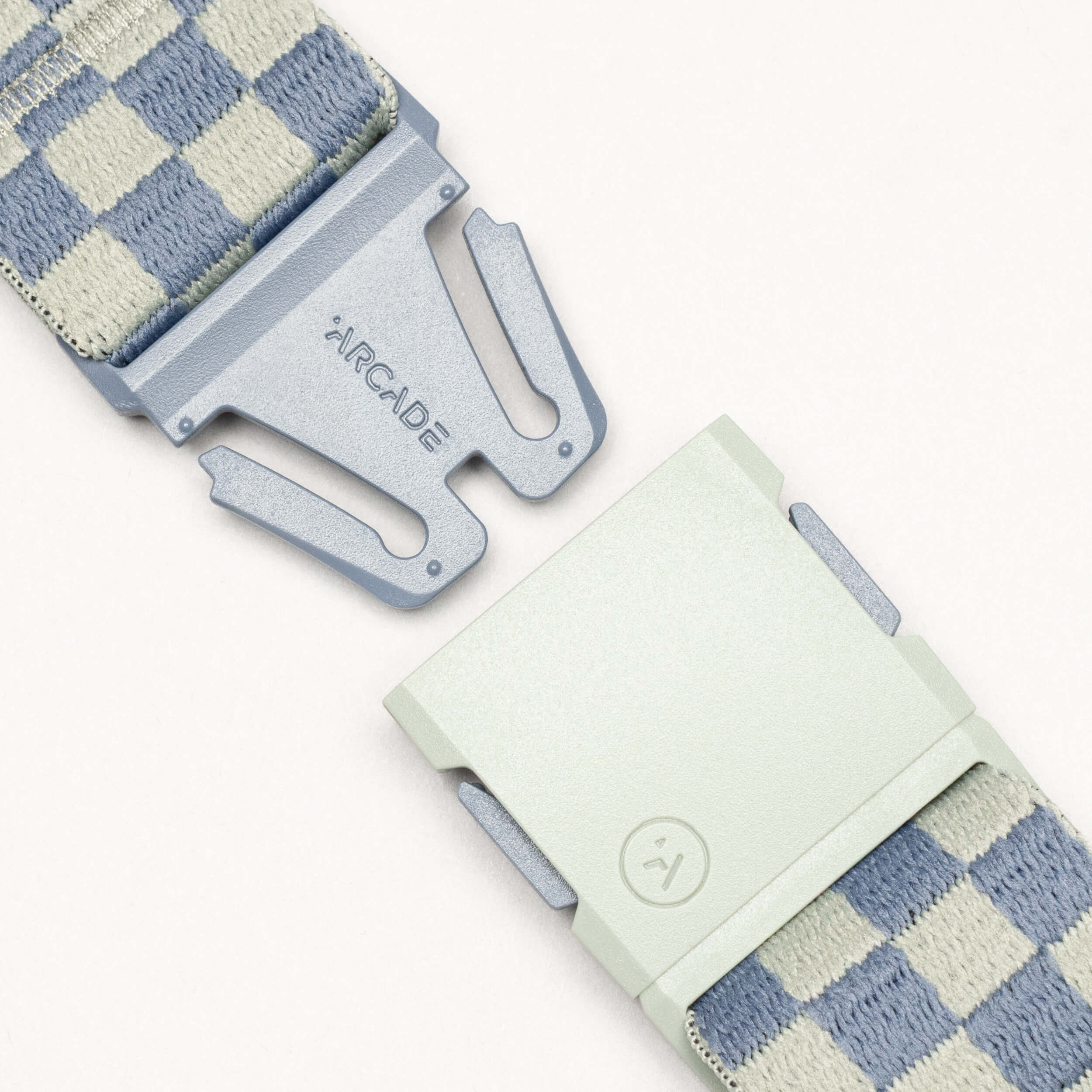 Close-up of the Arcade Belts Check: a light blue and beige checkered belt with performance stretch, designed for movement and comfort. The two-part clasp shows "ARCADE" on one side and a smiley face icon on the other.