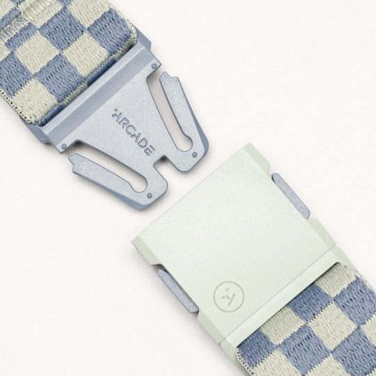 Close-up of the Arcade Belts Check: a light blue and beige checkered belt with performance stretch, designed for movement and comfort. The two-part clasp shows "ARCADE" on one side and a smiley face icon on the other.