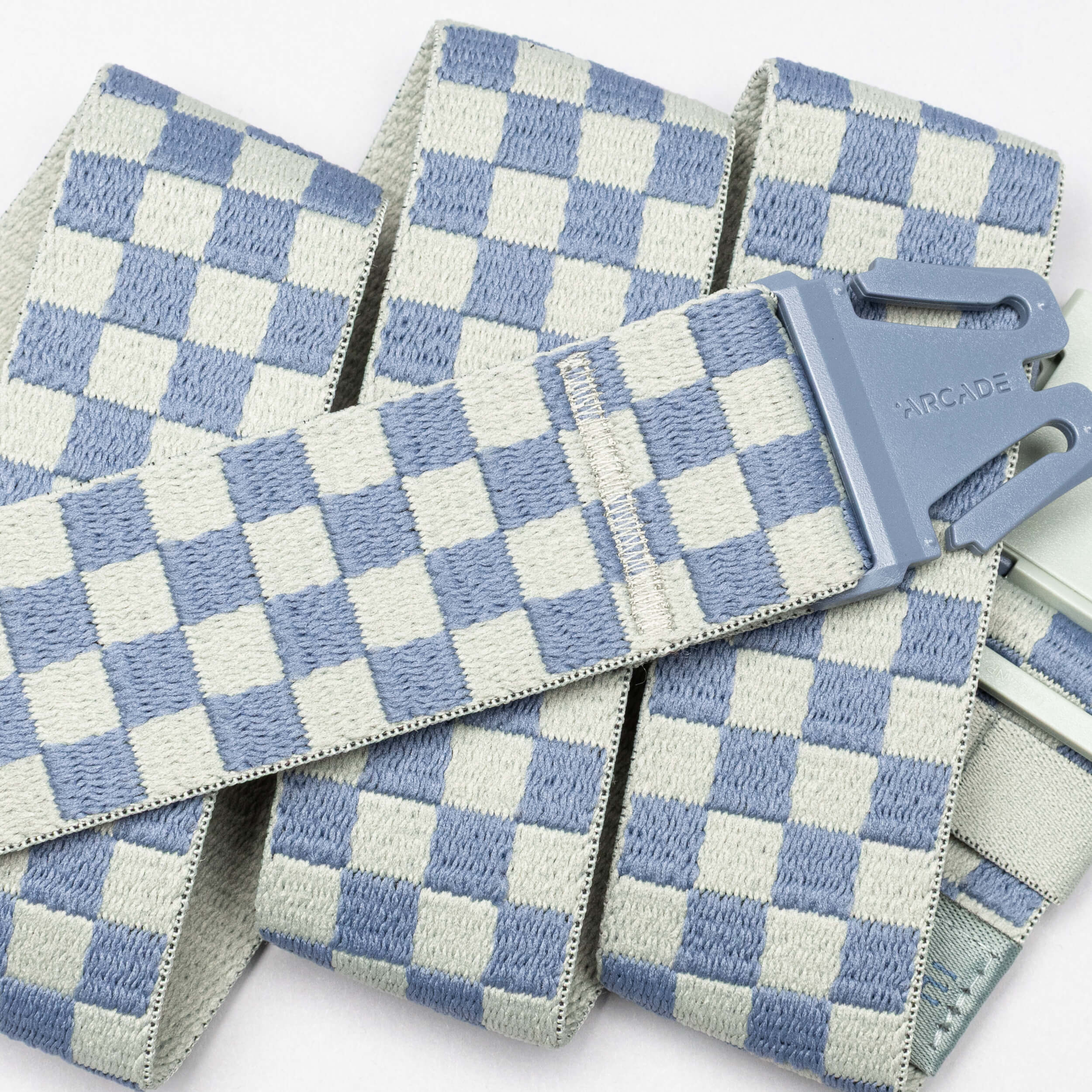 A close-up of the Arcade Belts "Check" shows its blue and white checkerboard pattern and gray buckle, featuring performance stretch for those who prioritize comfort in athletic wear.