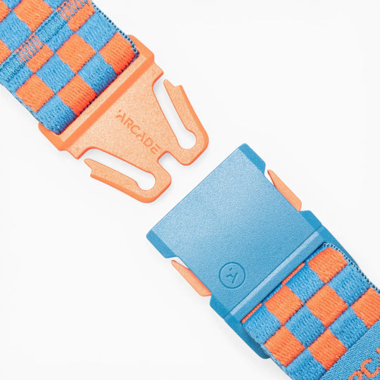 Here is the sentence rewritten using your product data: A close-up of the Arcade Belts "Check" belt buckle showcases a blue and orange checkerboard pattern, engineered for performance stretch. The partially unclasped buckle reveals "ARCADE" text on the orange segment. Designed for movement, this striking accessory stands out against a clean white background.
