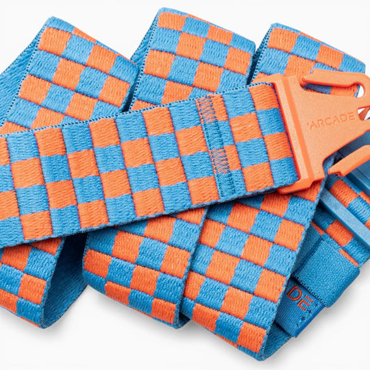 A folded Arcade Belts "Check" belt rests against a white background, showcasing its orange plastic buckle and a performance stretch elastic strap with a woven light blue and orange checkerboard pattern—designed for movement and comfort.