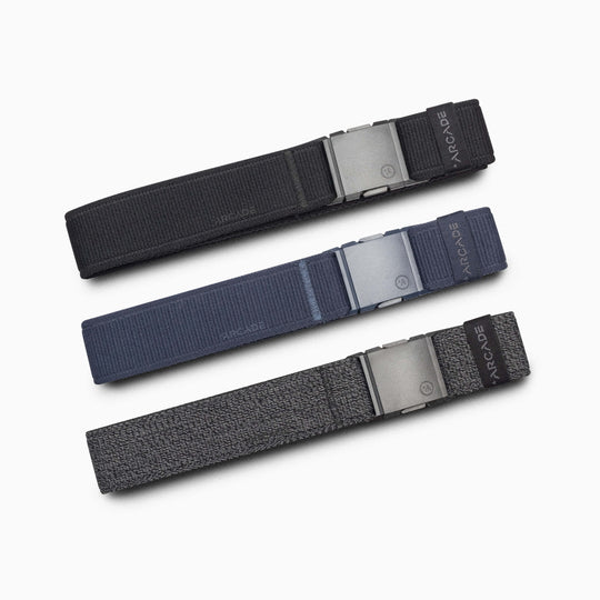 Three Atlas Kit fabric belts by Arcade Belts with metal buckles are laid out horizontally on a white background. The black, dark blue, and dark gray belts each display the “ARCADE” label near the buckle.