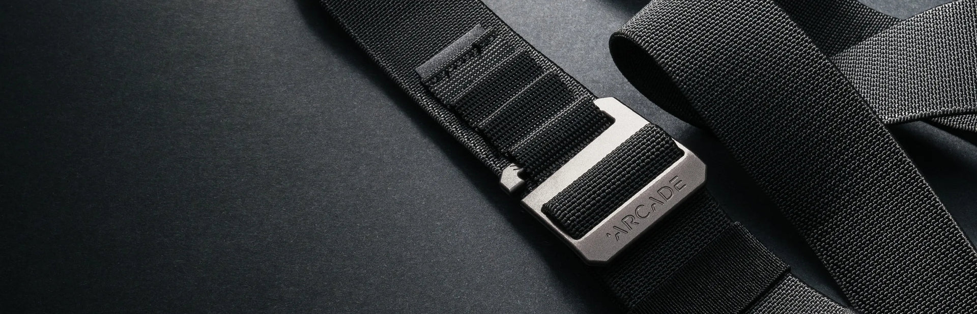 Close-up of a black fabric belt with a silver rectangular buckle engraved with the word ARCADE, resting on a dark, textured surface. The belt appears sturdy and modern.