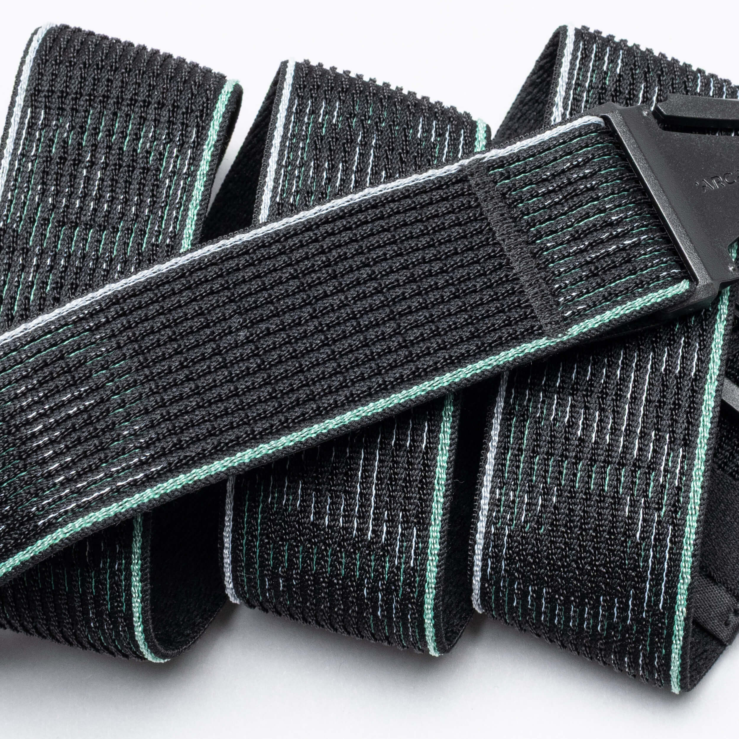 A close-up of the Arcade Belts Cubed: a black, textured fabric belt with mint green and white stitching, black plastic buckle, coiled comfortably on a white surface.
