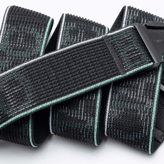 A close-up of the Arcade Belts Cubed: a black, textured fabric belt with mint green and white stitching, black plastic buckle, coiled comfortably on a white surface.