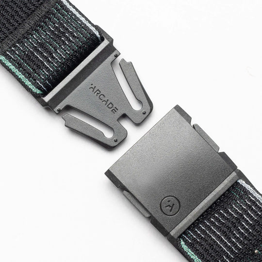 A close-up of the Cubed belt by Arcade Belts shows a black plastic buckle engraved with the brand, and both unfastened ends attached to a comfy black elastic strap featuring thin light blue stripes.