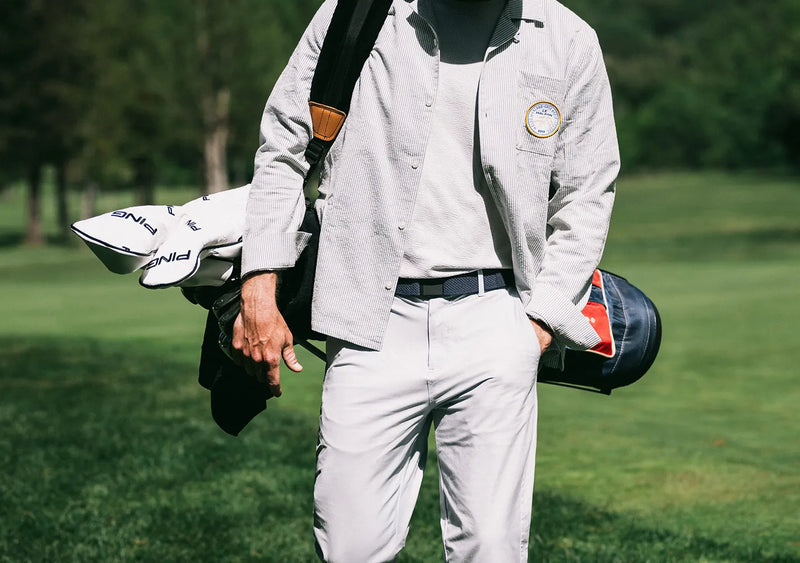 A person wearing light-colored pants and a button-up shirt carries a golf bag over their shoulder, standing on a green golf course with trees in the background.