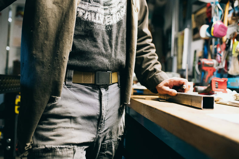 A person in casual work clothes stands at a workshop table, wearing a jacket and jeans secured with an elastic stretch belt, holding a metal square tube. Tools and equipment are visible in the background.
