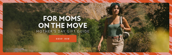 Woman hiking on a trail with a backpack, wearing a green tank top, beige shorts, and an Elastic Stretch Belt. The text reads For Moms on the Move - Mothers Day Gift Guide with a Shop Now button.