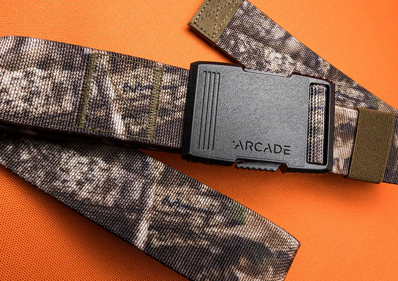 A camouflage-patterned belt with a black plastic buckle labeled ARCADE lies on an orange textured surface.