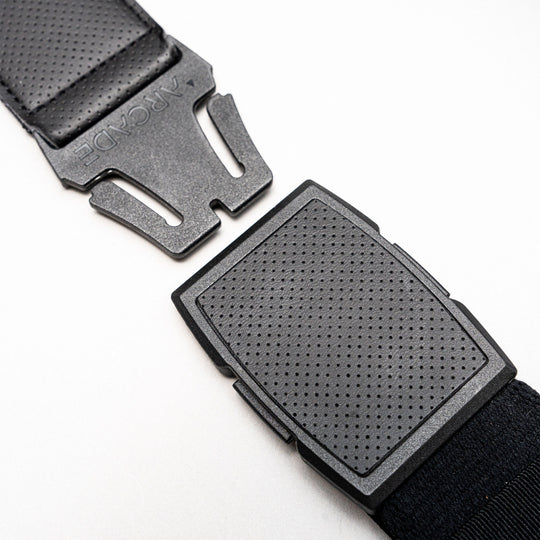 Close-up of a black plastic buckle on a strap, partially unclasped, lying on a white surface. The top piece is engraved with ARCADE, showcasing Arcade Belts’ Elevation stretch webbing design.