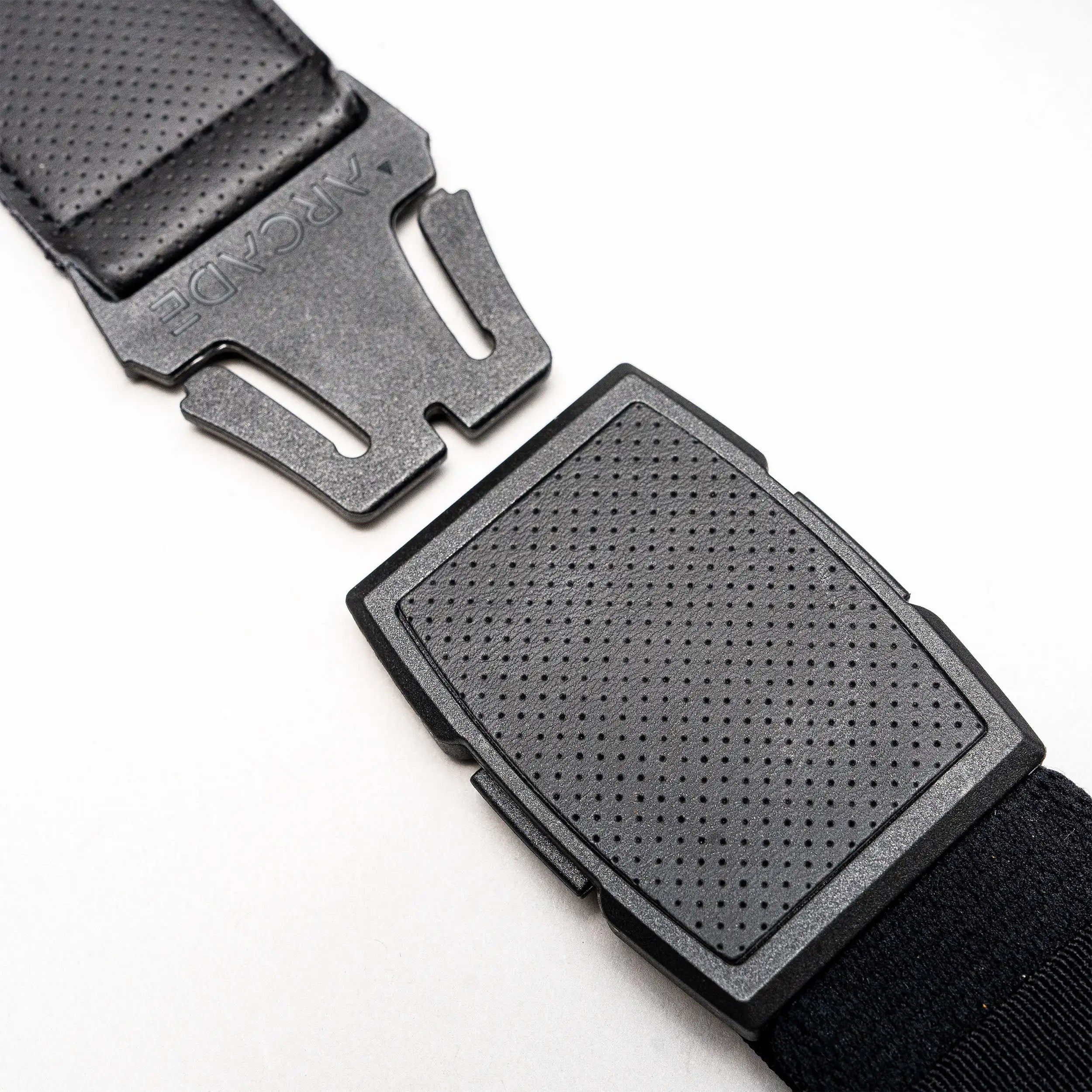 Close-up of a black plastic buckle on a strap, partially unclasped, lying on a white surface. The top piece is engraved with ARCADE, showcasing Arcade Belts’ Elevation stretch webbing design.