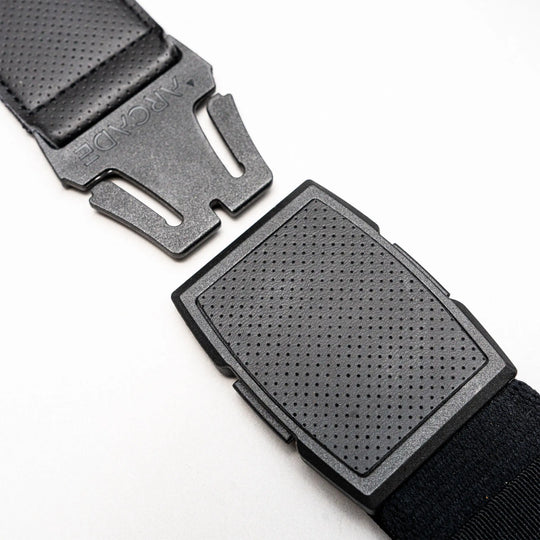 Close-up of a black plastic buckle on a strap, partially unclasped, lying on a white surface. The top piece is engraved with ARCADE, showcasing Arcade Belts’ Elevation stretch webbing design.