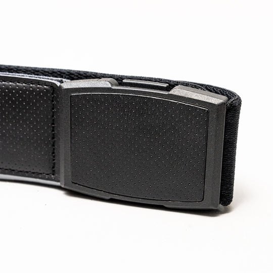 Close-up of the Elevation belt by Arcade Belts: a black, rectangular plastic buckle with a dotted pattern on a textured black stretch webbing strap combining fabric and leather, shown against a white background.