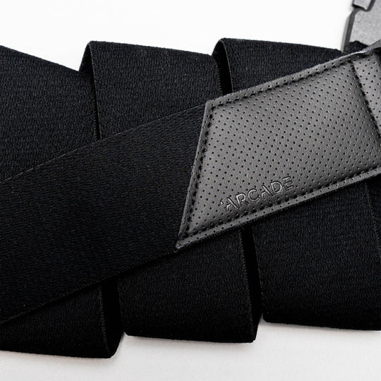 A close-up of Arcade Belts Elevation: a black travel belt with stretch webbing, a textured leather patch embossed with the logo, and a strong plastic buckle.