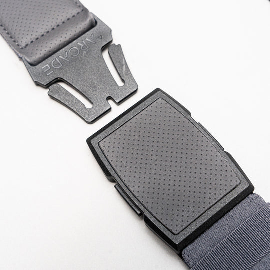 Close-up of a black, rectangular, textured plastic Elevation belt buckle with a perforated surface, partially open, attached to a dark gray Arcade Belts strap. This low profile belt is perfect for travel and everyday wear.