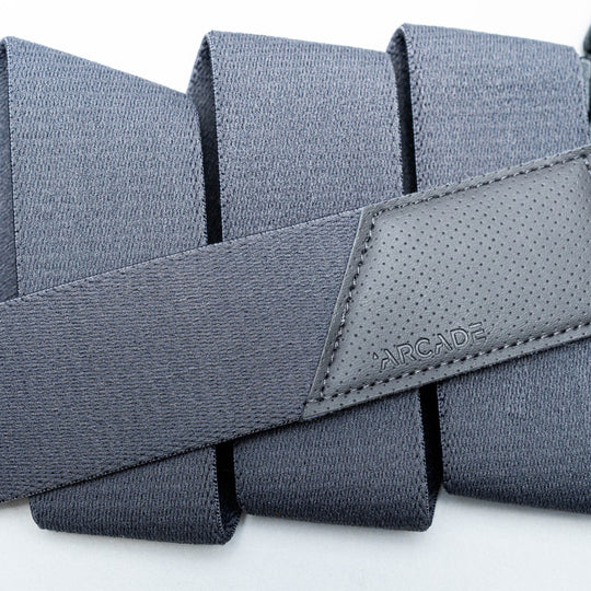 A close-up of the Elevation travel belt by Arcade Belts, featuring dark gray stretch webbing and a stitched leather patch embossed with the brand name, folded in neat layers on a light background.