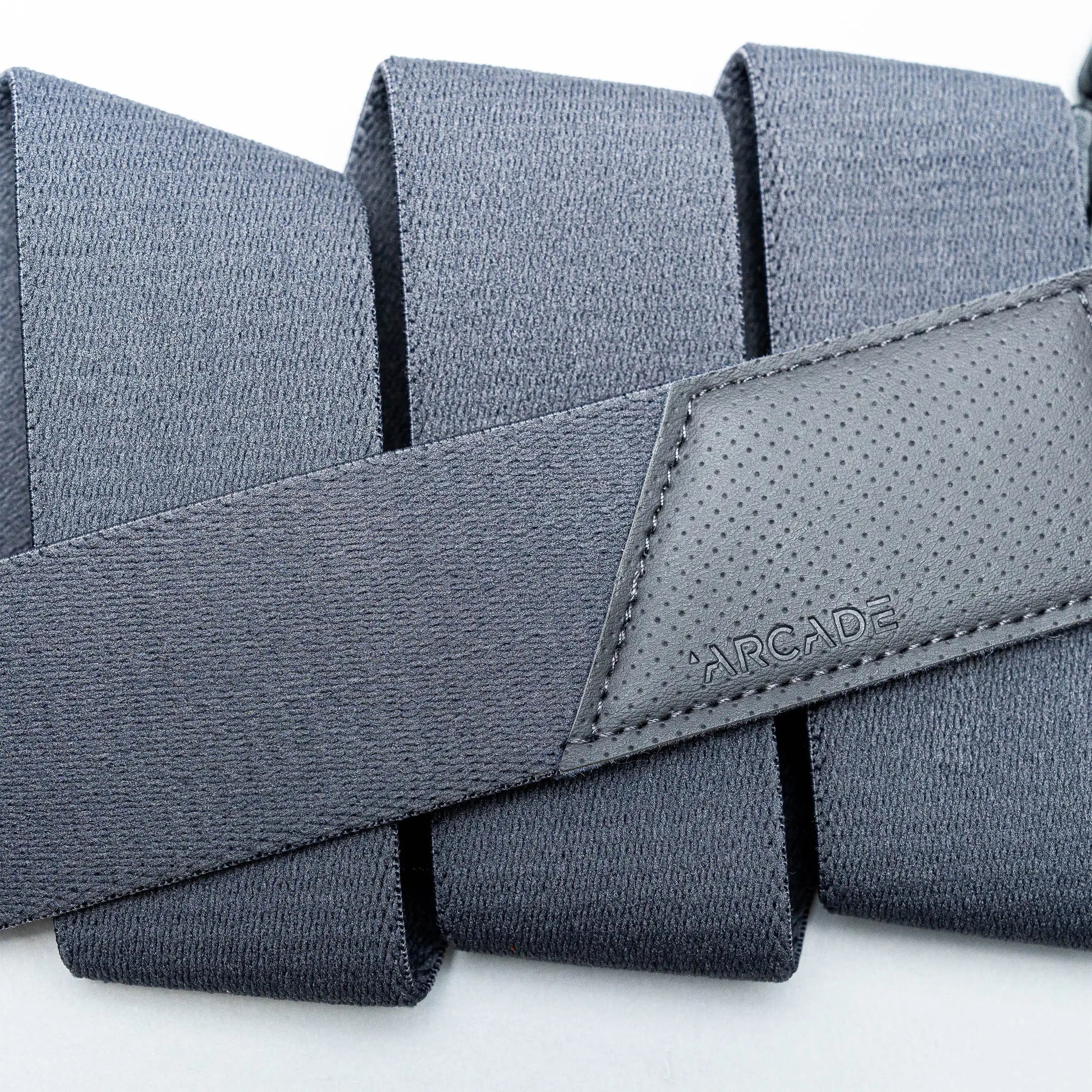 A close-up of the Elevation travel belt by Arcade Belts, featuring dark gray stretch webbing and a stitched leather patch embossed with the brand name, folded in neat layers on a light background.