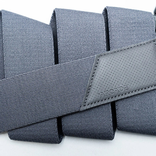 A close-up of the Elevation travel belt by Arcade Belts, featuring dark gray stretch webbing and a stitched leather patch embossed with the brand name, folded in neat layers on a light background.