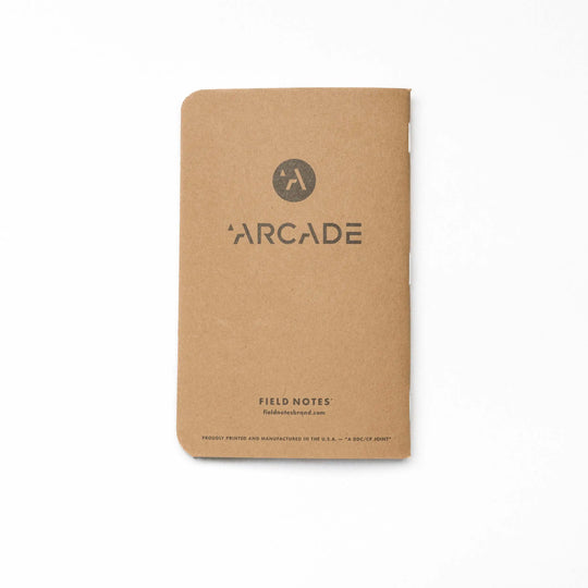 A brown, portable Field Notes notebook by Arcade Belts features a circular logo and "FIELD NOTES" plus the website on the cover. With durable construction, rounded corners, and a minimalist design, it’s ideal for on-the-go note-taking.
