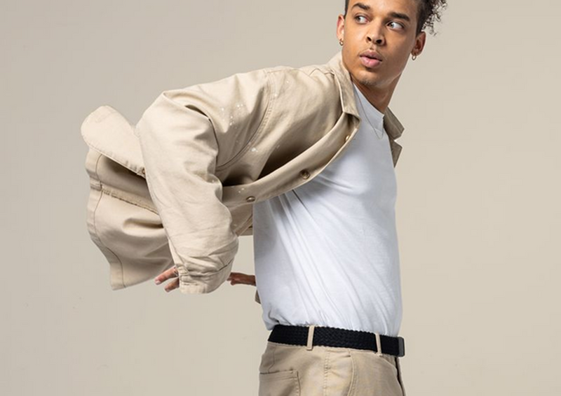 A young man wearing beige pants with a travel belt and a white t-shirt, putting on or taking off his beige button-up shirt while looking to the side, stands against a plain light background.
