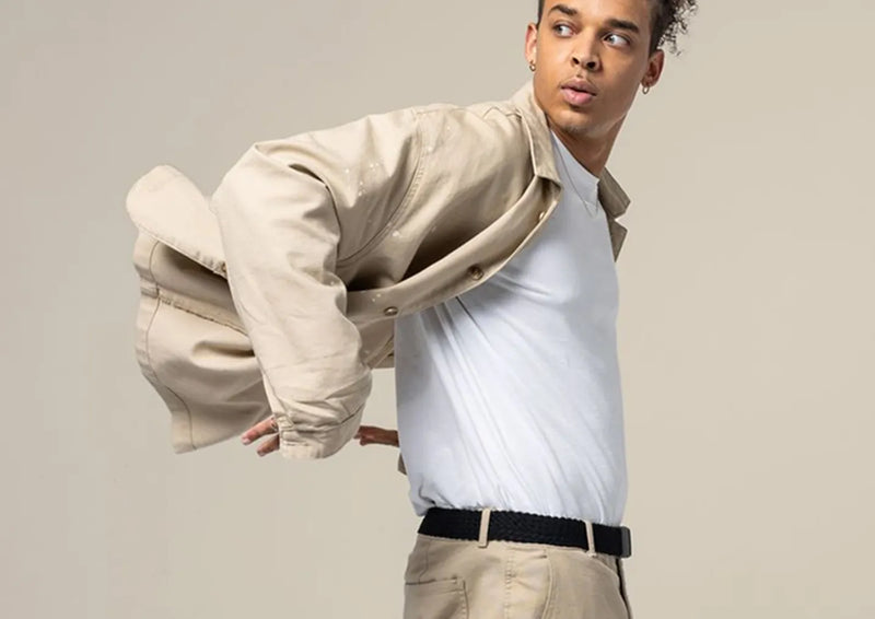 A young man wearing a white t-shirt, beige pants, and a beige jacket poses with one arm in his jacket, looking to the side against a neutral background.