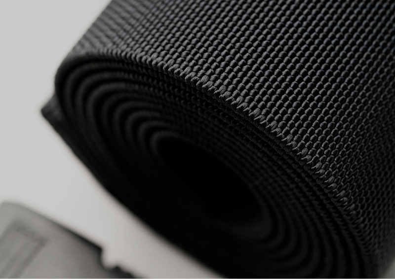 Close-up of a tightly rolled black woven fabric belt, showing its textured surface and spiral pattern. This Elastic Stretch Belt features a plastic buckle, while the blurred background highlights a dark out-of-focus object.