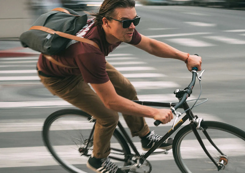A man wearing sunglasses, a maroon shirt, and brown pants rides a bicycle quickly through a city street, carrying a gray messenger bag over his shoulder. The background is blurred, showing motion.