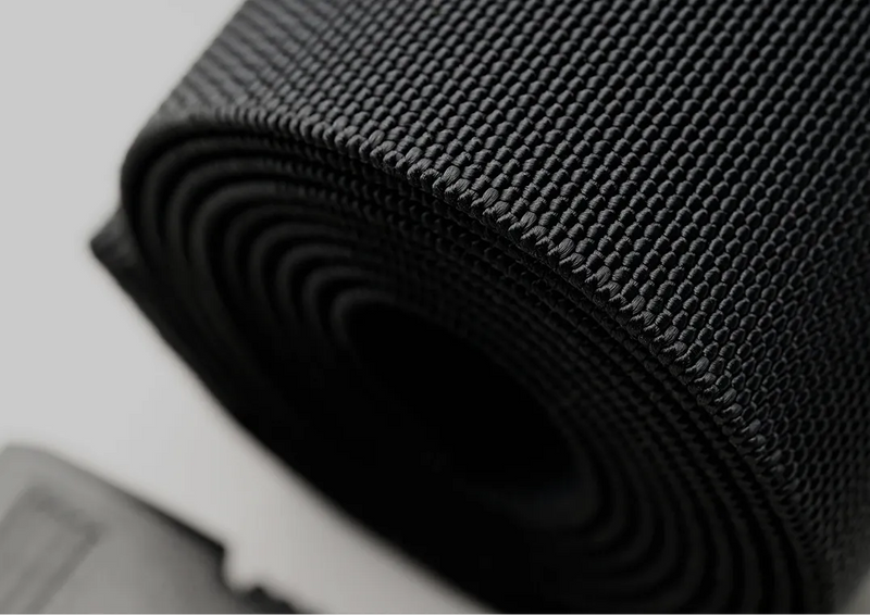 Close-up of a tightly rolled black nylon belt, showing the textured weave pattern. A small part of the black plastic buckle is visible in the lower left corner of the image.
