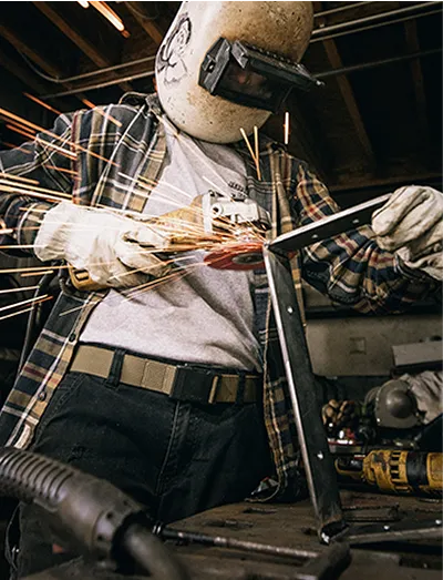 A person wearing safety gear, including gloves and a welding helmet, uses an angle grinder on metal, creating bright sparks in a workshop setting. Various tools and equipment are visible in the background.
