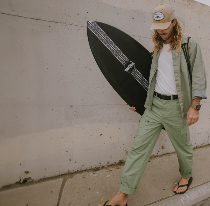 A person with long hair, wearing a beige cap, light green shirt and pants held up by an elastic stretch belt, and flip-flops, walks by a concrete wall carrying a black surfboard.