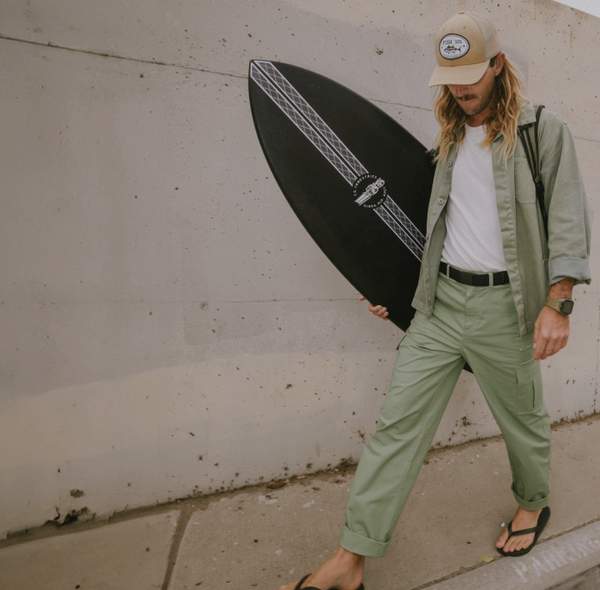 A person with long hair, wearing a beige cap, light green shirt and pants held up by an elastic stretch belt, and flip-flops, walks by a concrete wall carrying a black surfboard.