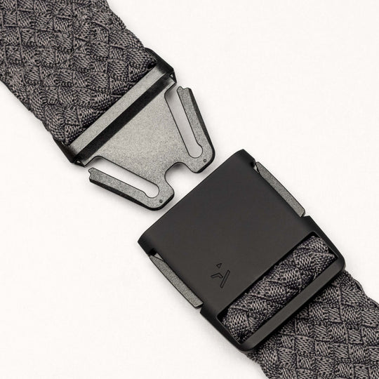 A close-up of the black metal buckle on the Arcade Belts Futureweave, featuring textured gray woven webbing, shown partially unfastened against a plain white background.