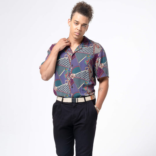 A person with curly hair stands before a white background, wearing a patterned short-sleeve shirt tucked into black pants with an Arcade Belts Lunar Kit buckle belt, one hand on their collar and the other in their pocket.