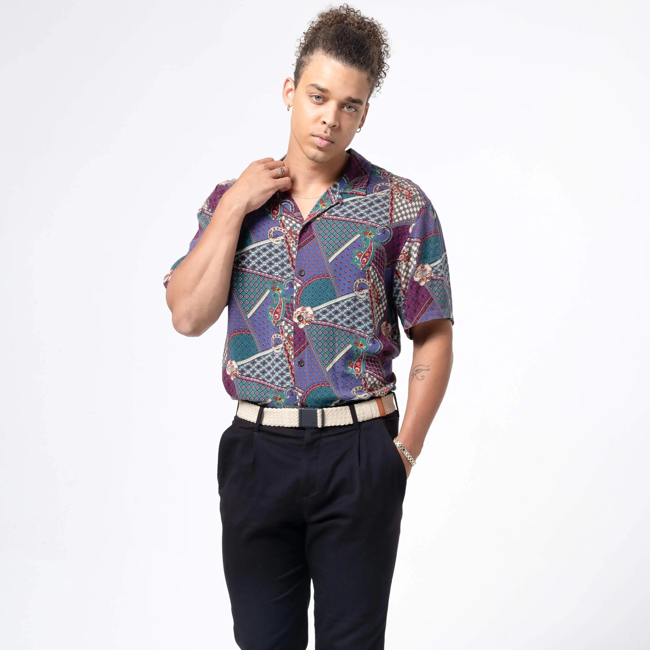 A person with curly hair stands before a white background, wearing a patterned short-sleeve shirt tucked into black pants with an Arcade Belts Lunar Kit buckle belt, one hand on their collar and the other in their pocket.