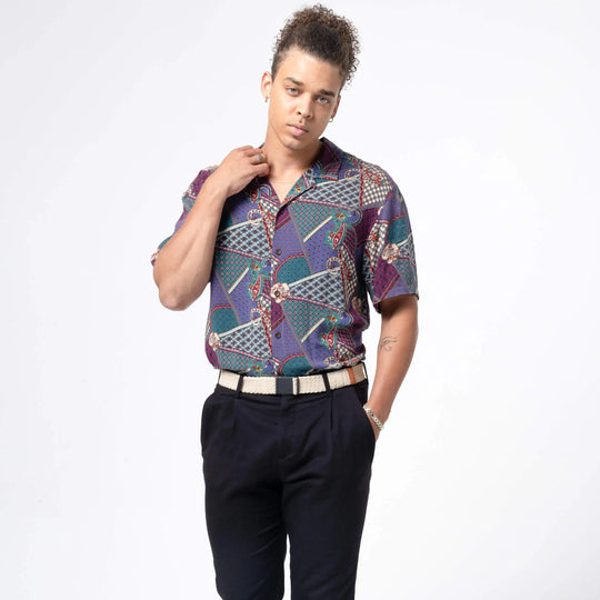 A person with curly hair stands before a white background, wearing a patterned short-sleeve shirt tucked into black pants with an Arcade Belts Lunar Kit buckle belt, one hand on their collar and the other in their pocket.