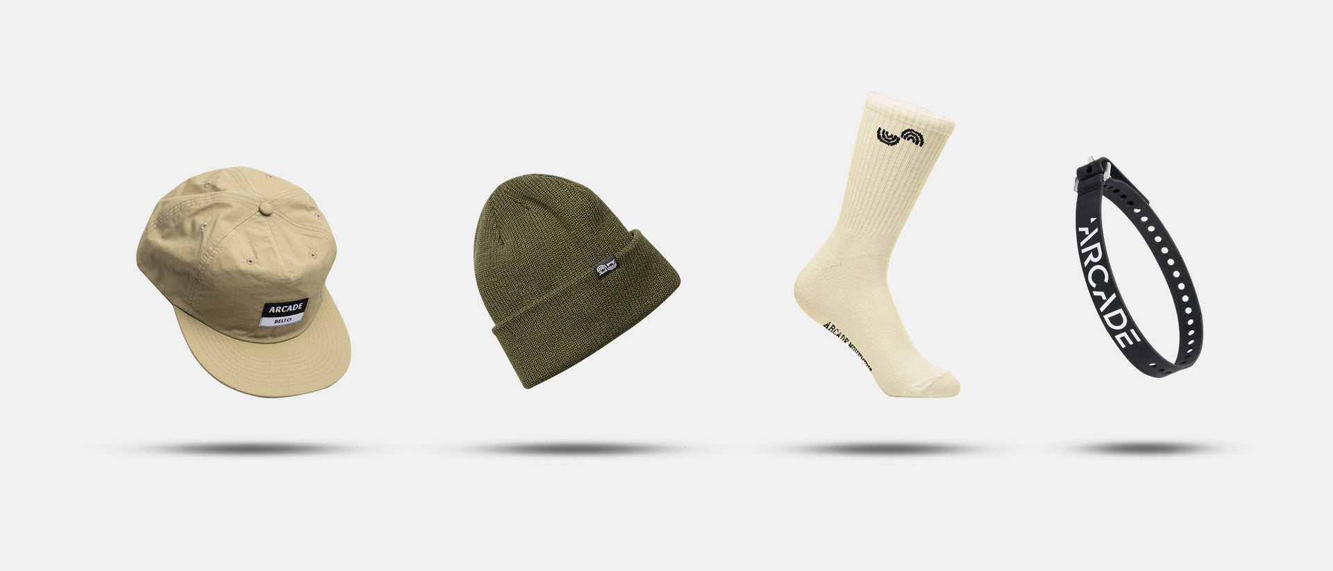 A beige cap, olive green knit beanie, cream-colored sock with a small logo, and a black Arcade Belt with plastic buckle branding—all floating with shadows on a white background. Perfect travel belts for everyday style.