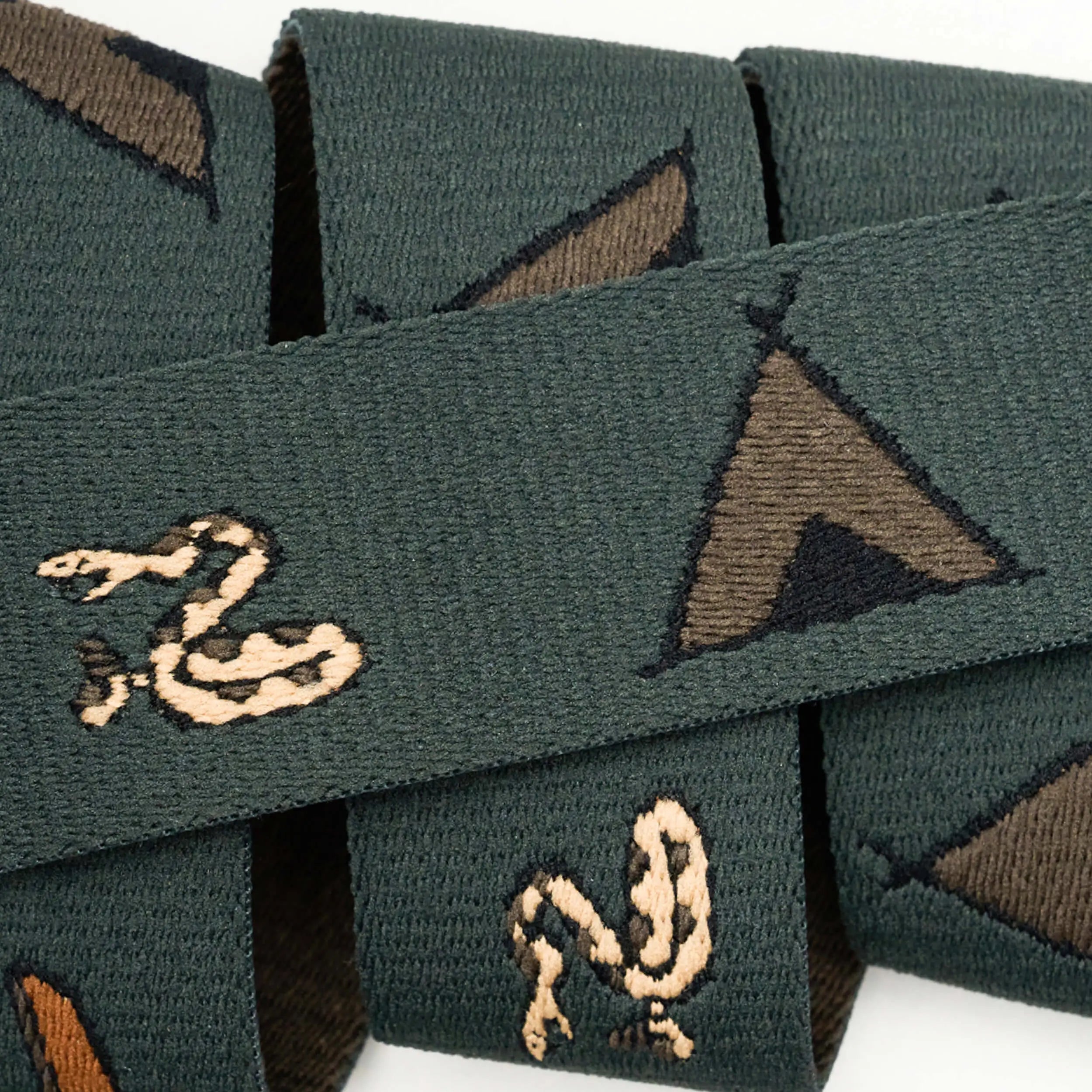 The Get Outside ribbon from Arcade Belts features green woven fabric with beige snakes and brown teepees, similar to Low Profile Belt designs. The ribbon is partially folded, creating overlapping layers.