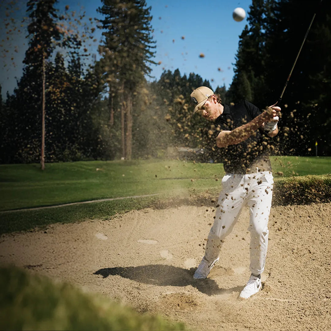 A golfer wearing a beige cap, black shirt, and white pants hits a ball out of a sand bunker on a sunny golf course, sending sand flying through the air with trees in the background.